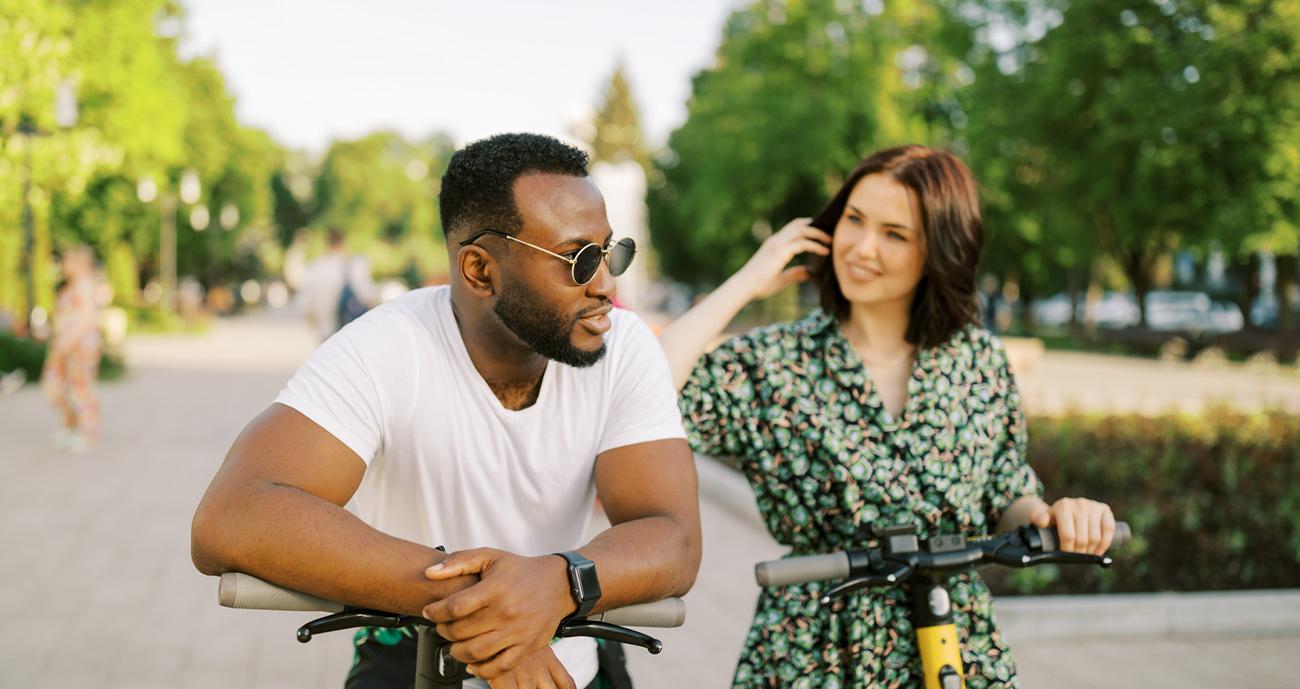 Man and woman together outside on an active date. The man is wearing a white T-shirt and the woman is wearing a patterned dress. She is looking at him.