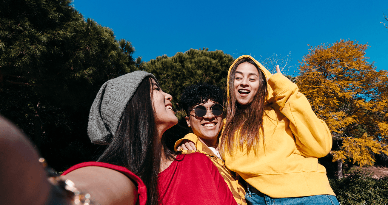 Three friends sitting together. One is wearing a yellow hoodie the other is wearing a red shirt. Another one is wearing sunglasses. It's a sunny day.