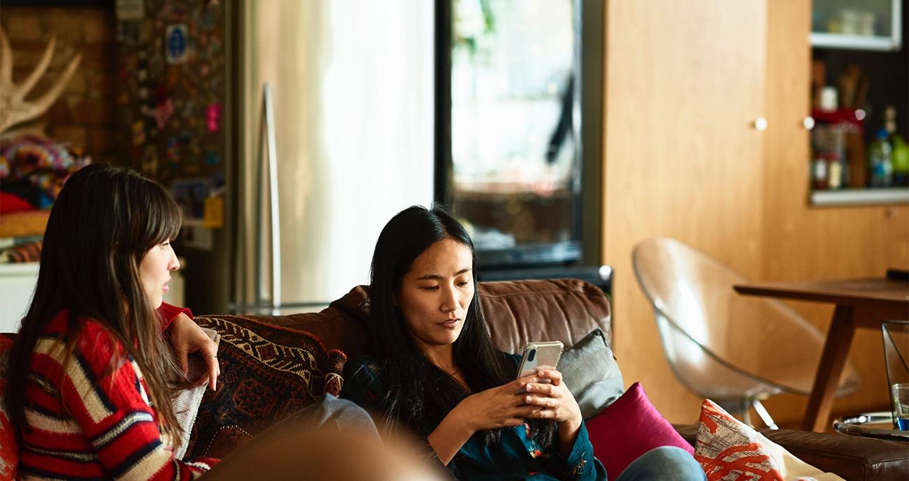A woman sits on a sofa next to a friend who is looking towards her. She is on her phone.