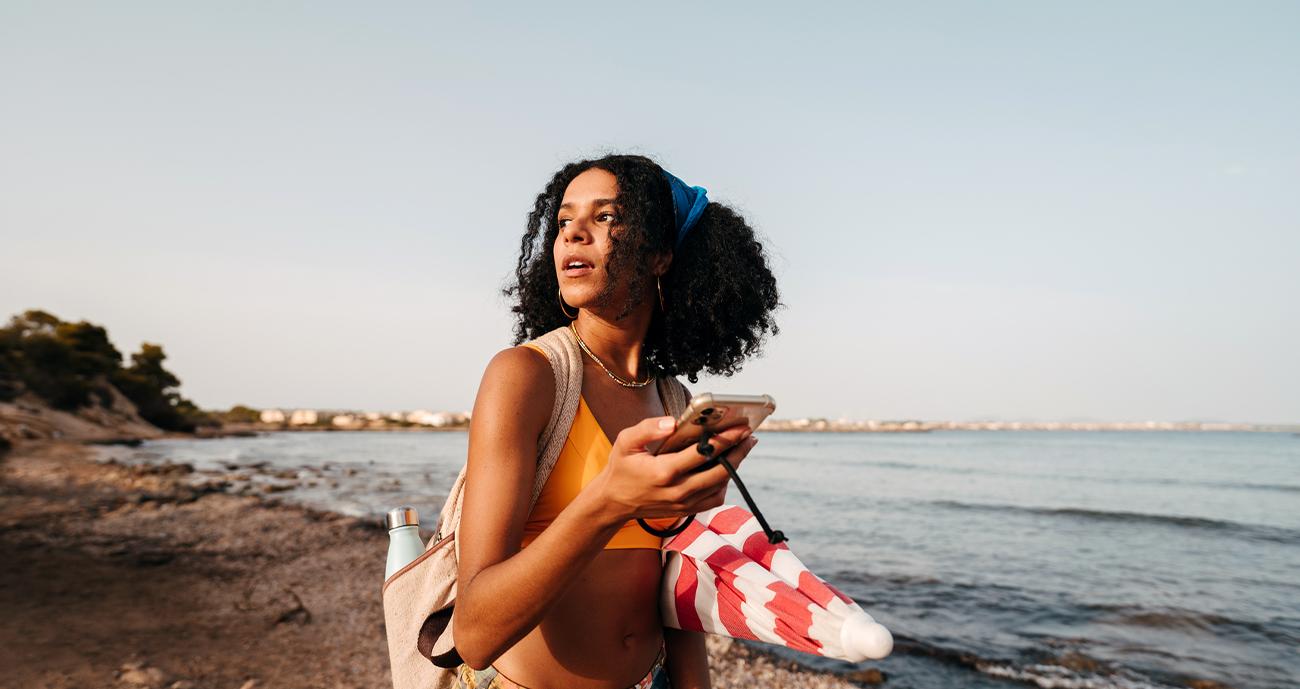 Woman on beach wearing a tank top, holding her phone and looking into the distance.