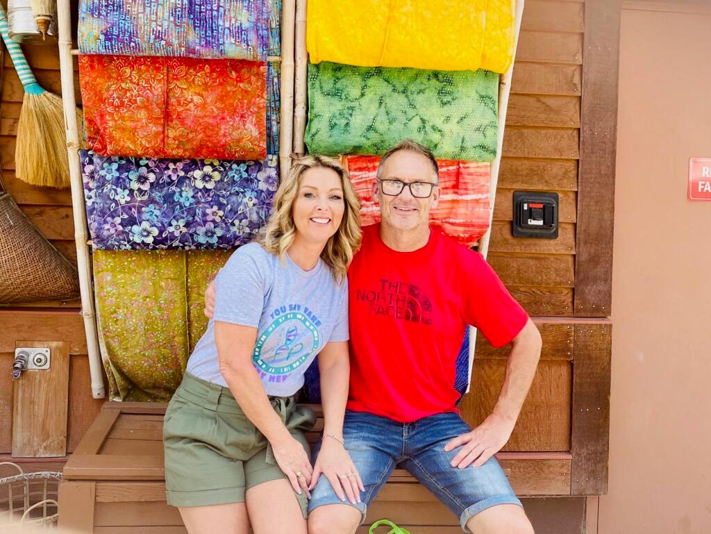 Helen and Chris are sitting together in front of a brightly coloured red background. Chris is wearing a bright red shirt and Helen is wearing a grey T-shirt and shorts.